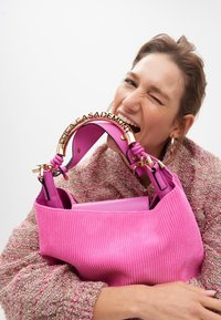 Una donna ammicca in modo giocoso mentre tiene in mano una vivace borsa fucsia con un manico alla moda. Indossa un maglione texturizzato e un orecchino a cerchio.