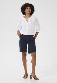 Vit, texturerad blus med rund halsringning och knytning, matchad med marinblå shorts. Modellen bär bruna klacksandaler med blockklack. Neutral bakgrund.