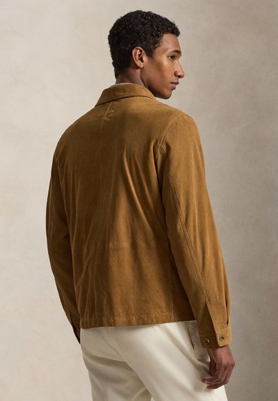 Uomo con giacca in suede marrone e pantaloni bianchi, in piedi di profilo contro uno sfondo beige uniforme.