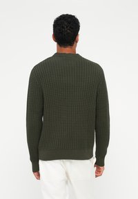 Maglione verde scuro e texture con un design a nido d'ape e polsini a costine. Presenta un collo a girocollo. Indossato con pantaloni di colore chiaro.