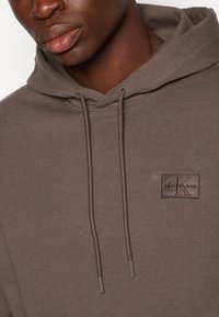 Felpa in cotone marrone con tasca a marsupio frontale, coulisse e un patch con il logo "Calvin Klein Jeans". Texture liscia.
