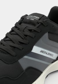 Black sneaker with grey accents, textured mesh and smooth rubber material. Features laces, logo on the tongue, and a contrasting white midsole.
