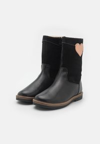 Par de botas negras de media caña con parte inferior de cuero, parte superior de ante, ribete marrón en la suela y parche beige en forma de corazón en el lado exterior de cada bota.