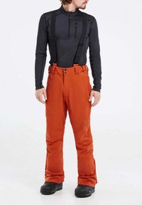 Oranga skidbyxor med justerbara hängslen, utrustade med knappar och fickor. Kombineras med en svart långärmad topp och svarta stövlar.