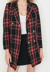 Femme portant un blazer à carreaux rouge, noir et blanc avec des boutons dorés sur un haut blanc et une jupe noire, debout devant un fond blanc.