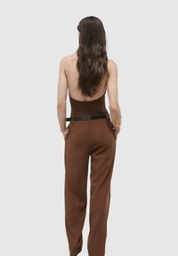 Combinaison brune sans dos avec un corsage ajusté, des pantalons à jambes larges et une ceinture noire mettant en valeur la taille. Texture lisse et design minimaliste.