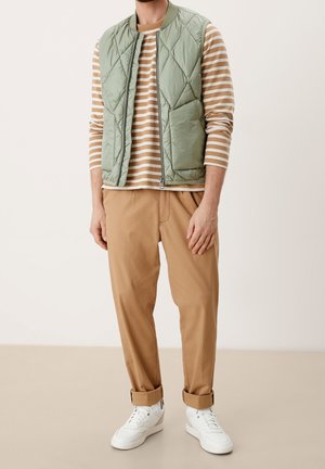 Man wearing light green quilted vest, tan striped long-sleeve shirt, camel pants with cuffs, and white sneakers standing on beige floor.