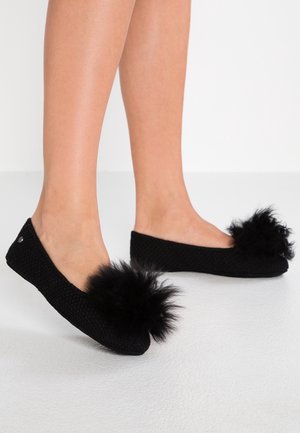 Beine in schwarzen gestrickten Ballerinas mit großen flauschigen Pompons an den Zehen, stehend auf hellem Boden vor weißem Hintergrund.