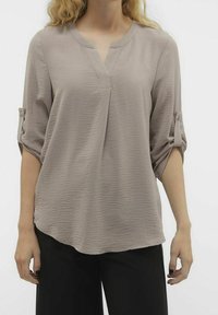 Blusa beige con texture, scollo a V, maniche a tre quarti arrotolabili e orlo curvato. Il materiale è leggero e leggermente sgualcito.