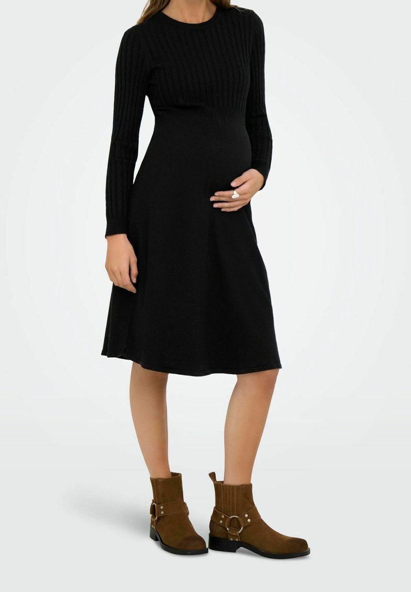 Femme enceinte portant une robe noire à manches longues et des bottines marron, tenant son ventre d'une main.