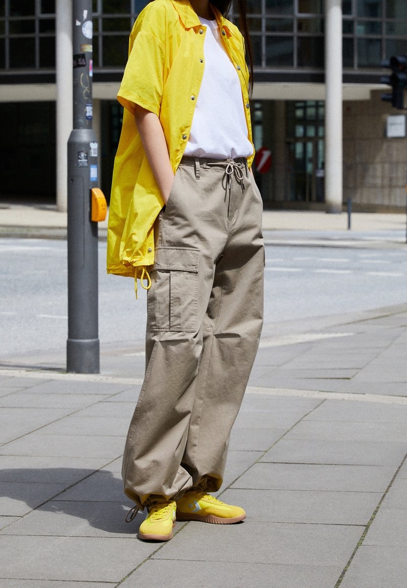 Personne portant un pantalon cargo beige surdimensionné, un t-shirt blanc, une veste jaune ouverte et des baskets jaunes debout sur un trottoir en ville.