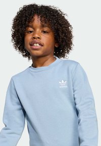 Svetlo modra majica s kapuco iz mehkega materiala. Ima okroglo ovratnik, dolge rokave in majhen bel logotip Adidas na prsih.