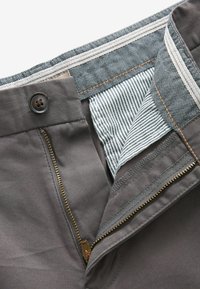 Harmaat housut, joissa on vetoketjukiinnitys ja erottuva sinivalkoraidallinen vuori. Mukana nappi ja kultainen vetoketjeyksityiskohta.