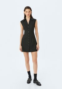 Schwarzes ärmelloses Kleid mit Knopfleiste, strukturiertem Kragen, figurbetonter Form und glattem Stoff. Getragen mit schwarzen Socken und Schuhen.