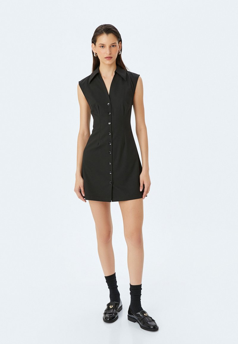 Schwarzes ärmelloses Kleid mit Knopfleiste, strukturiertem Kragen, figurbetonter Form und glattem Stoff. Getragen mit schwarzen Socken und Schuhen.