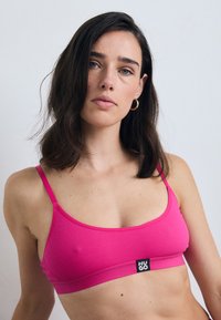 Rosa ribbet bralette med tynne stropper og et svart logomerke i midten. Enkelt design, myk tekstur og sømløs konstruksjon.