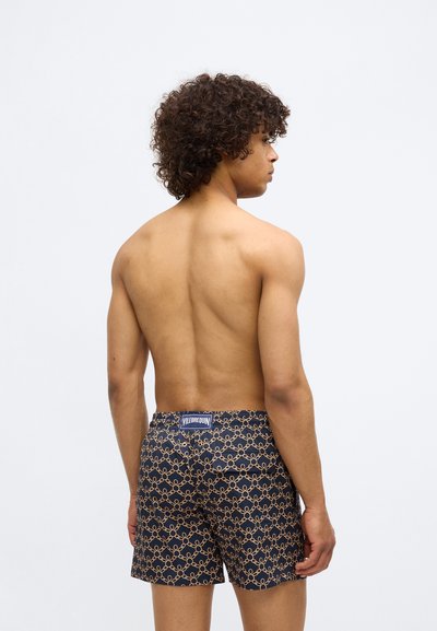 Vīrietis ar viļņainiem matiem, kaila auguma, ģērbies tumši zilos peldbiksēs ar zelta ķēdes rakstu un redzamu Vilebrequin markas etiķeti aizmugurē.