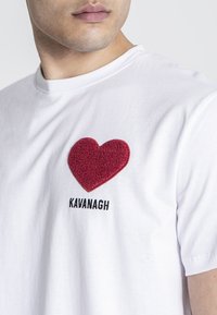 Camiseta blanca de algodón con un parche de corazón texturizado rojo en el pecho y el texto bordado en negro "KAVANAGH" debajo.