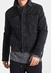 Svart denimjacka med långa ärmar, knäppning fram, två bröstfickor och klassisk krage. Buren över en strukturerad grå tröja.