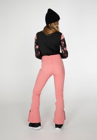 Pantalon de ski rose à jambes évasées avec fermetures éclair aux chevilles, associé à un haut noir à manches longues avec des manches à motifs floraux.