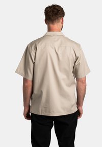 Chemise légère beige clair à manches courtes avec un col classique et une texture lisse, présentant un ourlet droit et des détails de couture minimaux.