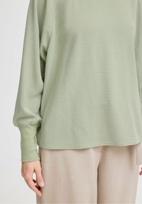 Maglione lungo a maniche verdi chiaro con una finitura texturizzata, vestibilità rilassata, polsini a coste e orlo arrotondato, abbinato a pantaloni beige.