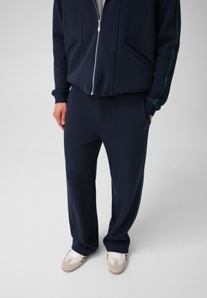 Mann trägt marineblaue Trainingshose mit Seitenstreifen und weiße Sneakers und steht vor einem einfarbigen Hintergrund.