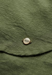 Tela verde oliva con una superficie texturada; presenta una insignia redonda en tono dorado con el logo de "Superdry" cosido en una solapa.