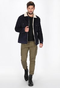 Marineblaues Cord-Jackett mit beigem Kunstpelzkragen, zwei Brusttaschen und vorderen Knöpfen, kombiniert mit olivgrünen Cargo-Hosen und schwarzen Schnürstiefeln.