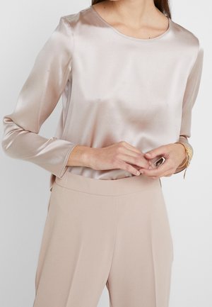 Kobieta ubrana w jasnoróżową satynową bluzkę z długim rękawem, wpuszczoną do beżowych spodni z wysokim stanem, z złotą bransoletką na nadgarstku.