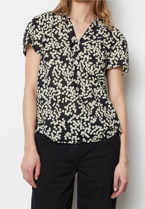 Vrouw draagt een zwart blouse met korte mouwen met een wit abstract stippenpatroon en zwarte broek, staand tegen een effen witte achtergrond.