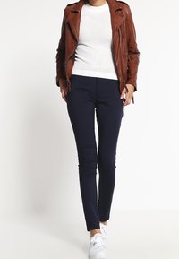Bruine leren motorjack, witte gestructureerde trui, marineblauwe slim-fit broek en witte sneakers. Focus op een slank ontwerp en contrasterende kleuren.
