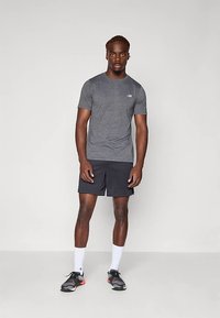 Homem em pé, vestindo uma camisa atlética cinza, calções pretos, meias brancas e sapatilhas de corrida pretas, contra um fundo claro e liso.