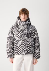 Giacca a vento con un motivo zigzag bianco e nero, colletto alto e texture spessa. Il design include una chiusura con zip e tasche laterali.
