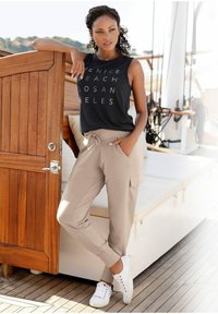 Schwarzes ärmelloses Oberteil mit weißem Text; beige schmal zulaufende Hose; weiße Sneakers; stehend neben einer Holztür auf einem Bootdeck mit Kissen.