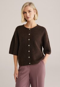 Barna kötött kardigán háromnegyedes ujjakkal, kerek nyakkivágással és világos színű gombokkal. Rózsaszín nadrág fölött viselve, puha tapintású anyagból.