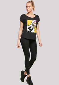 Svart t-skjorte med trykk av en tegneseriefigur på lys gul bakgrunn; kombinert med svarte leggings og svarte joggesko.