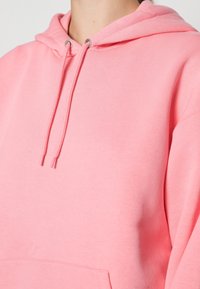 Sudadera de algodón rosa con capucha ajustable, bolsillo frontal y textura suave. Acentuada con ojales de metal en los cordones.