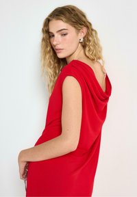 Abito rosso con collo a cappuccio, maniche corte, tessuto morbido, dettaglio drappeggiato sulla schiena, texture liscia, silhouette aderente. Indossato da una modella con capelli ricci.