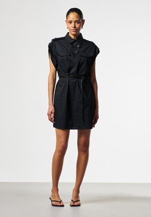 Frau trägt ein ärmelloses schwarzes Hemdkleid mit Gürtel und Fronttaschen sowie schwarzen Sandalen, stehend vor einem schlichten hellen Hintergrund.