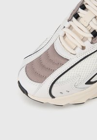 Αθλητικό παπούτσι με λευκό άνω μέρος από λείο δέρμα και δίχτυ, με ανάγλυφη ταμπέλα σε τόνο ταουπ, μαύρες λεπτομέρειες και στρογγυλή, μαλακή σόλα.