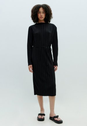 Schwarzes, plissiertes Midikleid mit langen Ärmeln und rundem Halsausschnitt, tailliert mit einem passenden Bindegürtel. Tragen mit klobigen schwarzen Sandalen.