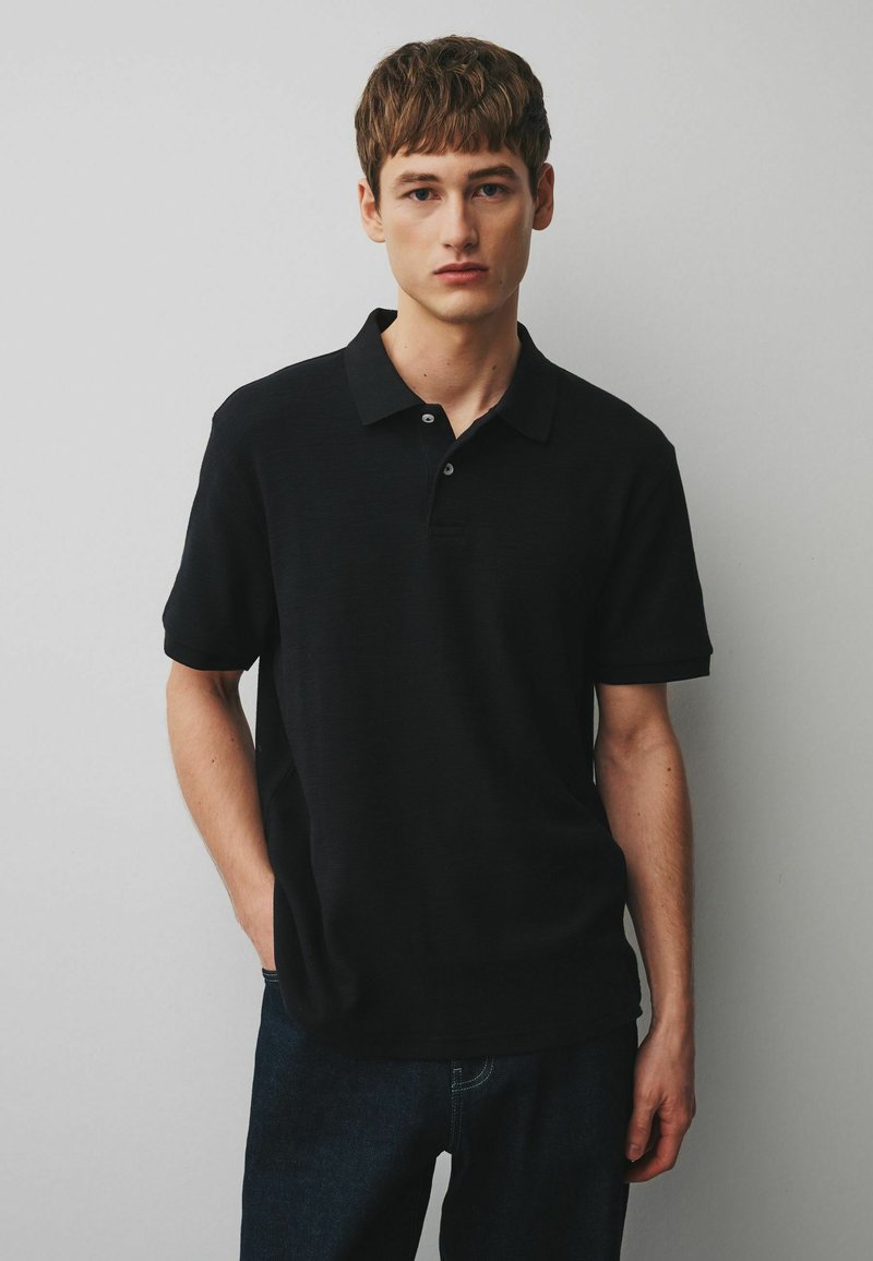 Fiatal, világos bőrű férfi rövid barna hajjal, egyszínű fekete rövid ujjú pólóingben és sötét nadrágban, egy egyszínű szürke fal előtt áll.