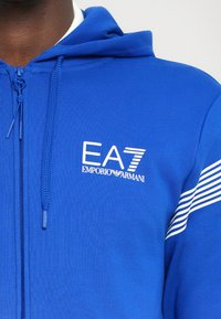 Blue zip-up hoodie made of soft fabric, featuring a white EA7 logo and two white stripes on the sleeves. Adjustable drawstring hood.