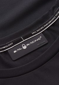 Sort beklædning med en glat tekstur, der har et halsmærke med teksten "SAIL RACING PERFORMANCE TECHNOLOGY" og størrelsen "MEDIUM" i hvid skrift.