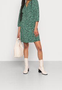 Vihreä kuvioitu mekko, jossa on kolme neljänneksen mittaiset hihat, yhdistetty valkoisiin polven korkeisiin nauhallisiin saappaisiin ja beigeen laukkuun. Sileä tekstuuri, vartalonmyötäinen malli.