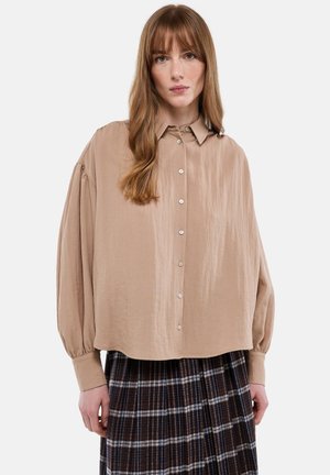 Femme aux longs cheveux châtain clair portant une chemise beige ample à boutons et une jupe écossaise marron, debout devant un fond uni.