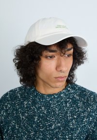 Boné branco com texto verde "MANTENHA A CALMA E BEBA MATCHA", usado com um suéter textura azul-petróleo, com gola redonda e padrão de tricô.