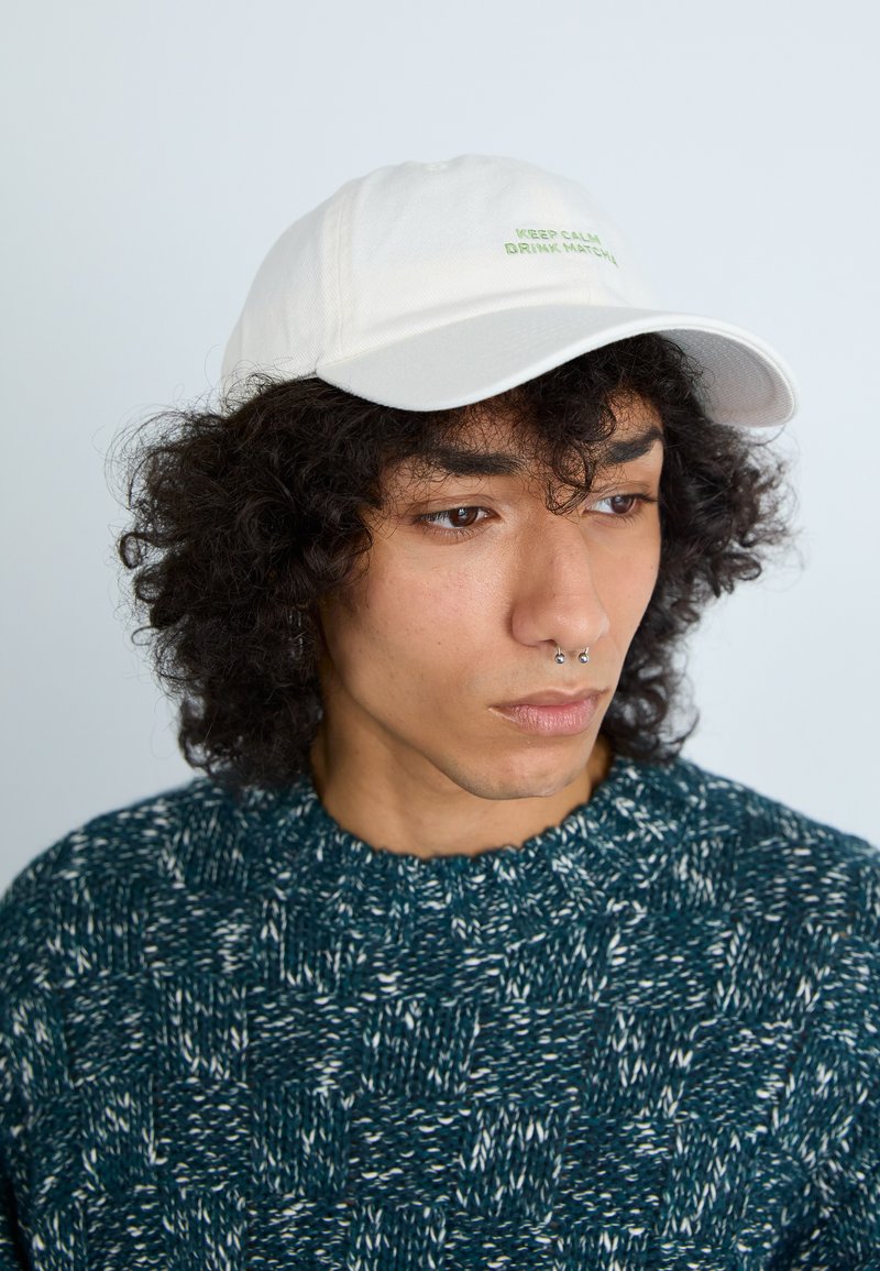 Boné branco com texto verde "MANTENHA A CALMA E BEBA MATCHA", usado com um suéter textura azul-petróleo, com gola redonda e padrão de tricô.