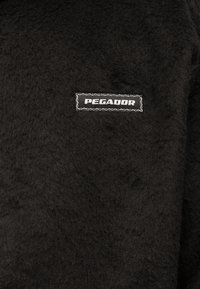 Vêtement en polaire noire à surface texturée. Une étiquette rectangulaire blanche avec l'inscription "PEGADOR" est bien visible.